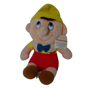 Op de afbeelding: Een pluche Pinocchio-pop met een gele hoed, zwart haar en een blauwe vlinderdas. De pop draagt een geel shirt, een rode korte broek en bruine schoenen. Een label op de pop leest "Pinocchio" en "Walt Disney Productions".