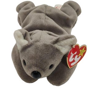 May include: A gray plush koala toy with a black nose and white inner ears. The toy has a red heart-shaped tag with gold lettering. The tag reads "Beanie Baby" and "Ty".