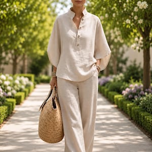 May include: A beige linen outfit consisting of a long-sleeved top with a mandarin collar and matching wide-leg pants. The outfit is paired with a woven straw tote bag. The model is walking on a stone path in a garden.