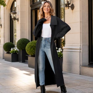 May include: A long-sleeved black duster cardigan worn over a white vest top and light blue denim jeans. The cardigan features a wide collar and an open front design that extends to the lower calf, paired with black ankle boots.