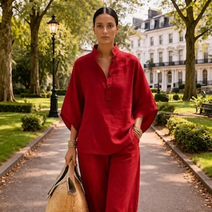 May include: A woman in a red linen outfit, including a top with a mandarin collar and button details, and matching wide-leg pants. She carries a woven tote bag and wears gold bracelets. The setting is a park with trees and a pathway.
