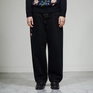 K&ouml;nnte beinhalten: Schwarze Jeans mit geradem Bein und floraler Stickerei in Rosa und Rot am linken Bein. Die N&auml;hte sind orange. Das Model tr&auml;gt schwarze Lederschuhe.