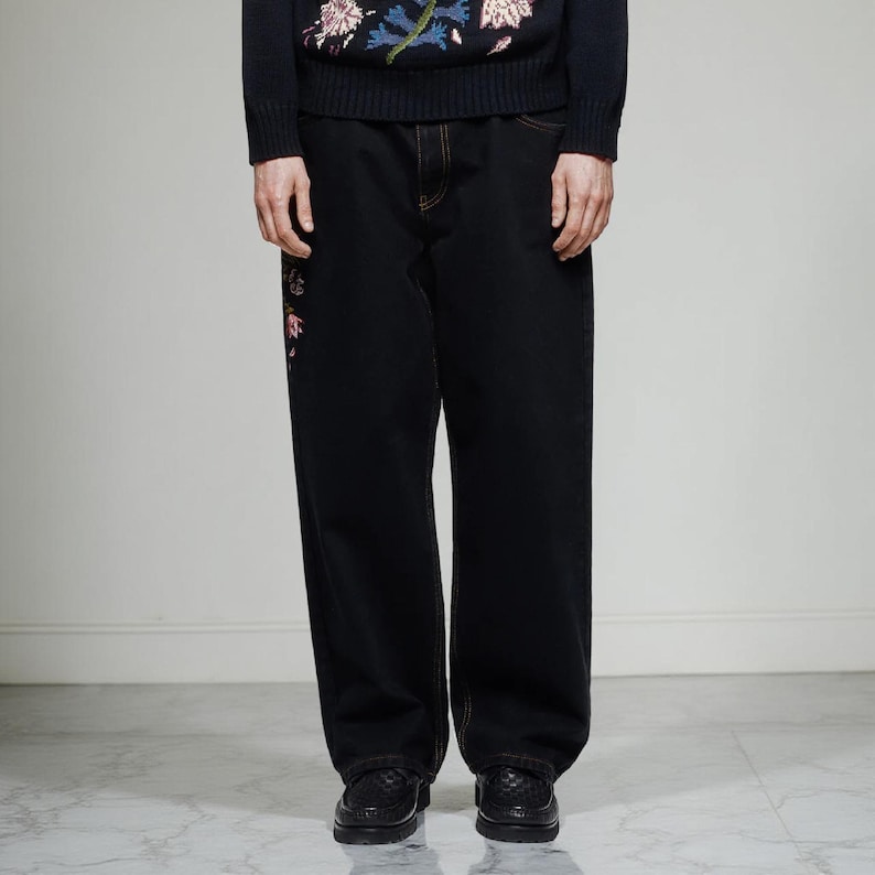 K&ouml;nnte beinhalten: Schwarze Jeans mit geradem Bein und floraler Stickerei in Rosa und Rot am linken Bein. Die N&auml;hte sind orange. Das Model tr&auml;gt schwarze Lederschuhe.