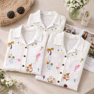 Puede incluir: Camisas blancas con botones con dise&ntilde;os bordados coloridos. Las camisas presentan palmeras, lunas, estrellas, flores y corazones. Las camisas son parte de un conjunto de ropa de dormir y se exhiben con un sombrero de paja, gafas de sol y una revista.
