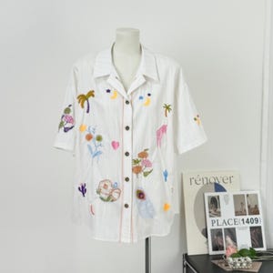 Puede incluir: Camisa blanca de manga corta con dise&ntilde;os bordados de flores coloridas, palmeras y lunas. La camisa tiene una botonadura frontal y un cuello. La camisa se exhibe en un maniqu&iacute;.