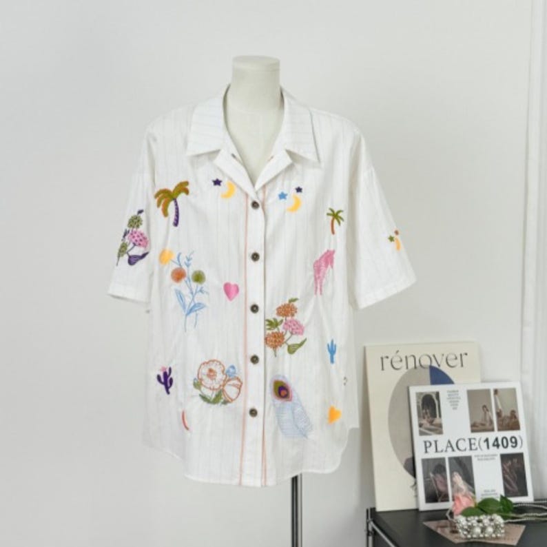 Puede incluir: Camisa blanca de manga corta con dise&ntilde;os bordados de flores coloridas, palmeras y lunas. La camisa tiene una botonadura frontal y un cuello. La camisa se exhibe en un maniqu&iacute;.