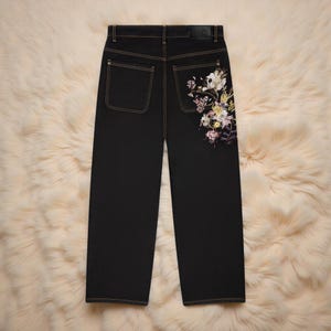 K&ouml;nnte beinhalten: Schwarze Jeans mit geradem Bein. Die rechte Ges&auml;&szlig;tasche ist mit floralen Stickereien in Wei&szlig;, Rosa, Gelb und Lila verziert. Die Jeans haben ein klassisches F&uuml;nf-Taschen-Design mit beigen N&auml;hten.