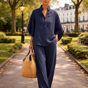 May include: A navy blue linen two-piece outfit, consisting of a top with a mandarin collar and wide-leg pants. The top has a button-down front and short sleeves. A woven straw tote bag is carried. The outfit is worn outdoors on a paved walkway.