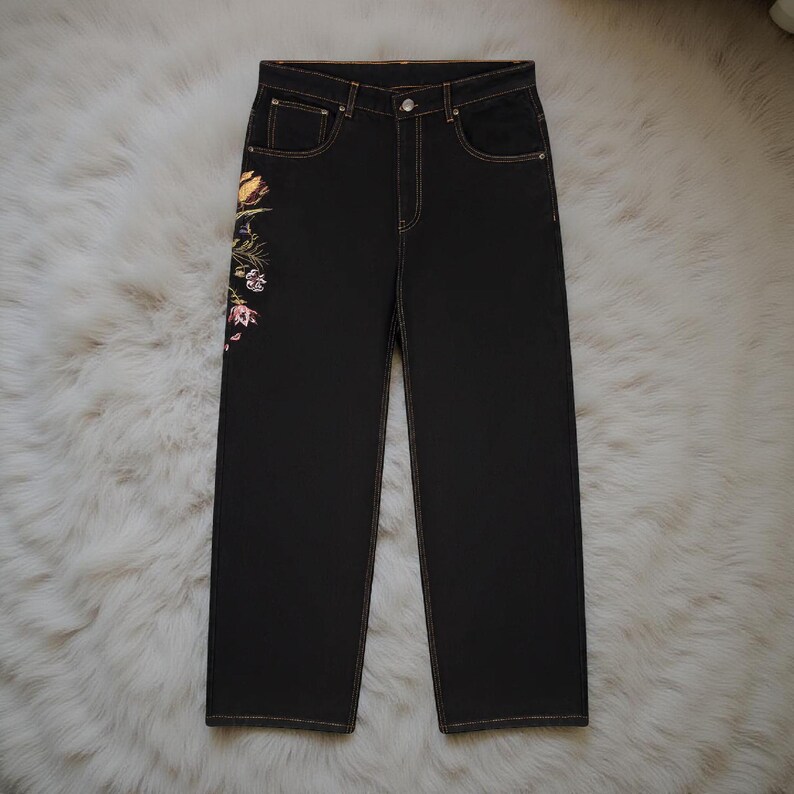 K&ouml;nnte beinhalten: Dunkelblaue Jeans mit geradem Bein. Die Jeans haben eine Blumenstickerei in Gelb, Rosa und Gr&uuml;n am linken Bein. Die N&auml;hte sind in einem kontrastierenden Beigeton gehalten.