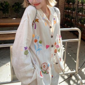 Puede incluir: Camisa crema con botones y dise&ntilde;os bordados coloridos de flores, frutas y celestiales. La camisa tiene mangas largas y un corte relajado. El bordado incluye una palmera, flores y una luna creciente.