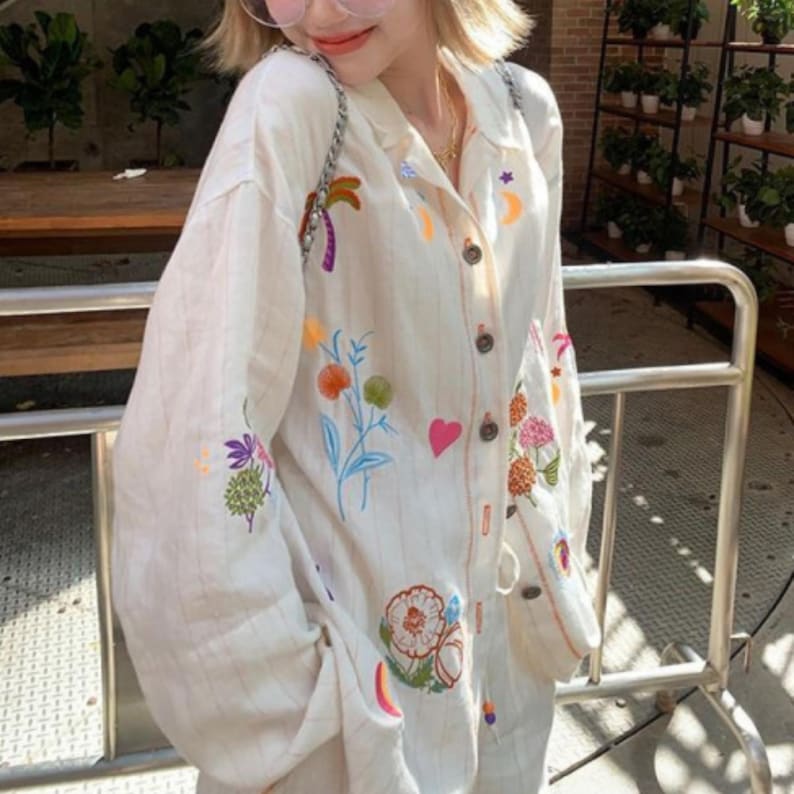 Puede incluir: Camisa crema con botones y dise&ntilde;os bordados coloridos de flores, frutas y celestiales. La camisa tiene mangas largas y un corte relajado. El bordado incluye una palmera, flores y una luna creciente.