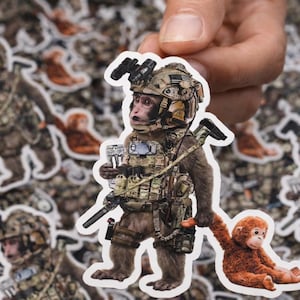 May include: A sticker featuring a monkey in military gear, holding a weapon and a small orange monkey. The sticker has a white border and is held by a hand. The background is filled with similar stickers.