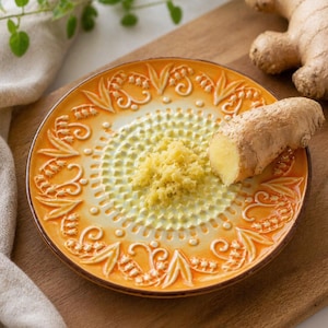 May include: A ceramic ginger grater with a yellow and orange floral design. The grater has a textured surface and is filled with grated ginger. A piece of fresh ginger sits on the side, with a whole ginger root in the background.