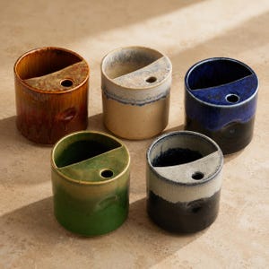 May include: Five ceramic vases in various colors: brown, tan, blue, green, and black. Each vase is cylindrical with a diagonal cut across the top and a small hole. The vases have a glossy finish and are arranged on a neutral surface.