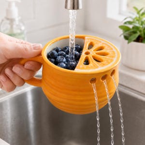 May include: A bright orange ceramic berry colander shaped like a citrus fruit with a handle and a built-in strainer lid. The colander is filled with fresh blueberries being rinsed under a stream of water, which drains through holes.