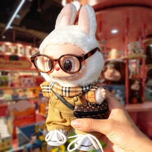 Puede incluir: Un peluche de conejito con gafas, una bufanda a cuadros y un mono de pana. El conejito tiene un peque&ntilde;o bolso negro y zapatillas blancas. El juguete tiene una capucha de piel blanca con orejas internas rosas.