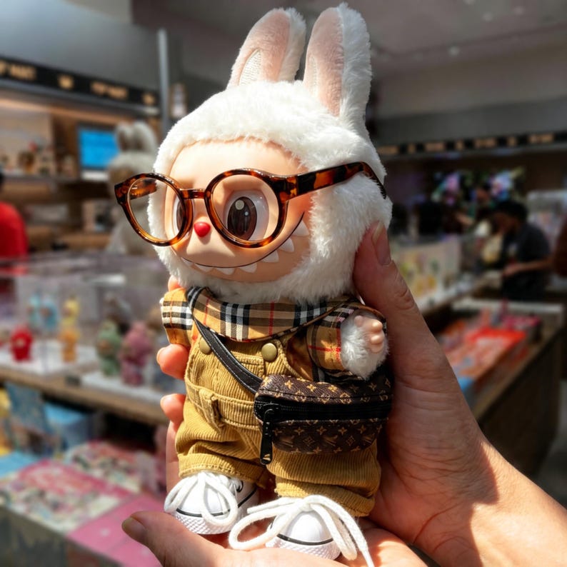Puede incluir: Una figura de peluche de conejo con gafas de carey, una bufanda a cuadros, un peto de pana y un peque&ntilde;o bolso cruzado. La figura tiene pelaje blanco, orejas grandes y zapatillas blancas. La figura est&aacute; siendo sostenida en la mano de una persona.