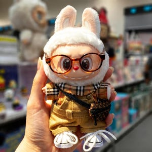 Puede incluir: Una figura de peluche con forma de conejo que lleva un gorro de piel blanco, gafas con montura marr&oacute;n y una camisa a cuadros. La figura viste un mono color canela, una peque&ntilde;a bolsa negra y zapatillas blancas. El juguete est&aacute; en la mano de una persona.