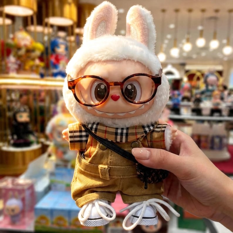 Puede incluir: Una figura de peluche de un conejo con una capucha de piel blanca con orejas rosas, gafas de carey y una bufanda a cuadros. La figura viste un mono marr&oacute;n, un bolso negro y zapatillas blancas.