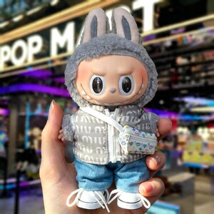 Pu&ograve; includere: Una statuetta in vinile da collezione di un personaggio che indossa un cappello con orecchie da coniglio grigio, un gilet imbottito grigio, jeans blu e scarpe da ginnastica bianche. La statuetta ha una piccola borsa bianca con un motivo colorato. La statuetta ha un'espressione facciale unica.