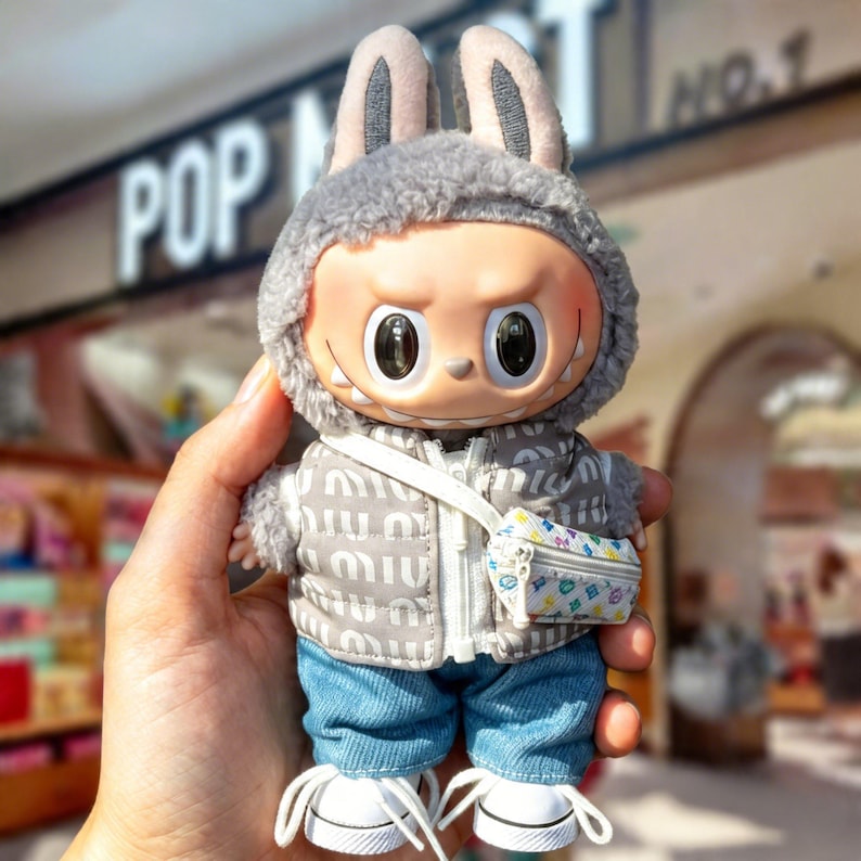 Pu&ograve; includere: Una statuetta in vinile da collezione di un personaggio con una felpa con cappuccio grigia con orecchie da coniglio, una giacca trapuntata, jeans blu e scarpe da ginnastica bianche. La statuetta ha una piccola borsa fantasia e un'espressione severa. Lo sfondo ha la parola "POP".