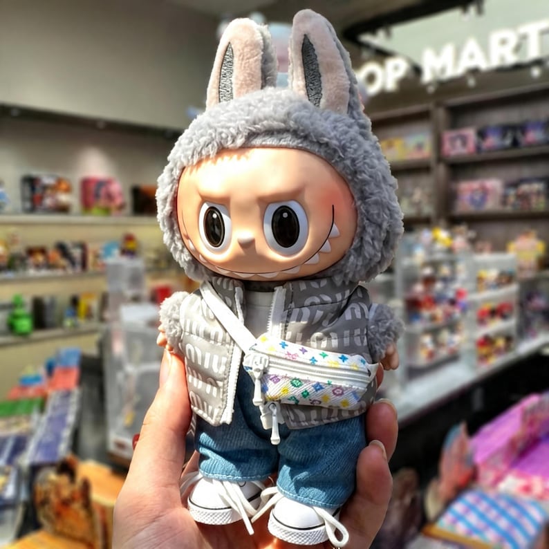 Pu&ograve; includere: Una statuetta in vinile da collezione di un personaggio che indossa un cappuccio grigio con orecchie da coniglio, un gilet imbottito grigio, jeans blu e scarpe da ginnastica bianche. La statuetta ha una piccola borsa a tracolla con un motivo colorato. La statuetta ha un'espressione severa.