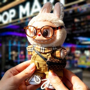 Puede incluir: Figura de conejo coleccionable con gafas, bufanda a cuadros, peto de pana y un peque&ntilde;o bolso. La figura tiene una cara sonriente y lleva zapatillas blancas. El fondo est&aacute; borroso, con las palabras "POP MART" visibles.