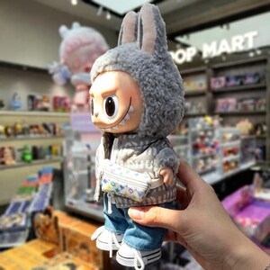 Pu&ograve; includere: Una statuetta in vinile di un personaggio con cappello e giacca grigi a forma di coniglio, jeans blu e scarpe da ginnastica bianche. La statuetta ha un sorriso malizioso e una piccola borsa fantasia. La statuetta si trova in un negozio con l'insegna "POP MART".