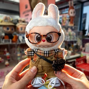 Puede incluir: Una figura de peluche con forma de conejo que lleva gafas, una camisa a cuadros, un peto de pana y zapatillas blancas. El conejo tiene un peque&ntilde;o bolso negro y un sombrero blanco y peludo con orejas interiores rosas. La figura est&aacute; sostenida en manos.