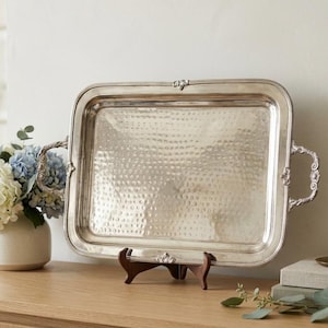 May include: A rectangular silver-toned metal serving tray with a hammered texture on the base and ornate, curved handles on each side. The tray features a raised border with decorative detailing and is displayed upright on a small wooden stand.