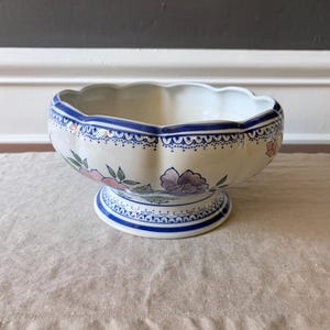 Puede incluir: Un cuenco de cerámica de color crema con borde festoneado y base de pedestal. El exterior presenta un patrón floral pintado a mano en tonos rosa, morado y verde, acentuado por un borde decorativo azul y blanco en la parte superior y la base.