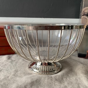 Puede incluir: Un cuenco de vidrio transparente con borde y base de metal plateado. Los lados del cuenco están revestidos con un diseño decorativo de alambre plateado. El cuenco está sobre una superficie gris.