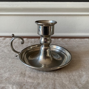 May include: A silver-colored candlestick holder with a round base and a handle. The holder has a cup-shaped top to hold a candle. The handle is curved and has a decorative swirl at the end. The candlestick holder is made of metal and has a shiny finish.