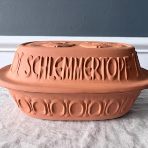 May include: An oval terracotta clay cooking pot with a lid. The lid features the embossed text SCHLEMMERTOPF and two stylized bird figures. The base of the pot is decorated with a repeating circular pattern around the exterior side.