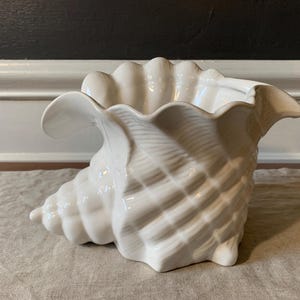 May include: A white ceramic seashell-shaped planter. The planter has a glossy finish and detailed ridges, with a wide opening and flared edges. The seashell design is a decorative accent for coastal or nautical-themed decor.