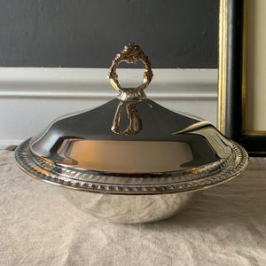 May include: A silver serving dish with a lid and ornate handle. The dish has a round base and a decorative rim. The lid is domed and reflects the surrounding environment. The handle is gold-colored and has a detailed, floral design.