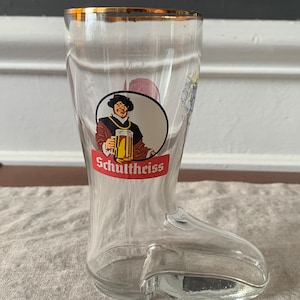 May include: Clear glass boot-shaped beer glass with a gold rim. The glass features a circular emblem with a man holding a beer stein and the word "Schultheiss" in red. The glass is on a neutral-colored surface.