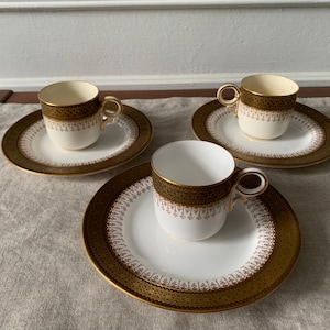 May include: Three matching teacups and saucers. The cups and saucers are white with a gold and brown decorative border. The cups have a gold rim and handle. The saucers are oval-shaped.