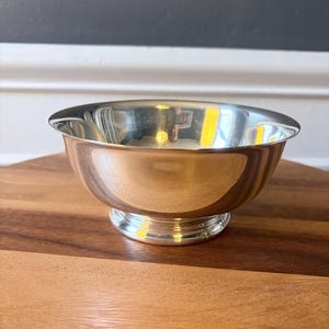 May include: A polished silver-toned bowl with a smooth, reflective surface and a wide, circular rim. The bowl features a rounded body that tapers down to a sturdy, elevated circular base with subtle horizontal ridged detailing.