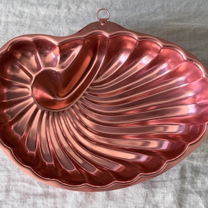 May include: A metallic copper-colored baking mold shaped like a large seashell. The mold features a spiraled, ribbed design with deep grooves radiating from the center and includes a small circular metal loop at the top for hanging purposes.