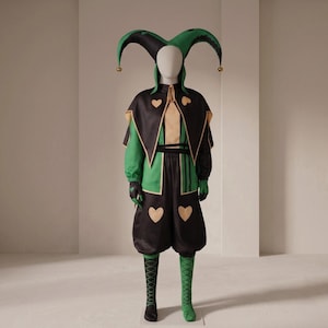 May include: A jester's costume on a mannequin, featuring a black and green colour scheme with heart motifs. The hat has three points with gold baubles. The outfit includes a cape, tunic, and bloomers. The costume is designed for a theatrical performance.