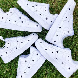 May include: White sneakers with black star patterns. The shoes have a classic design with a low-top silhouette. The star pattern is evenly distributed across the upper and sides of the shoes. The sneakers are arranged on a green grassy surface.