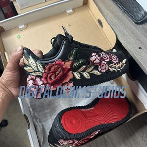 May include: A black sneaker featuring intricate floral embroidery in shades of red, pink, and green along the side. The shoe has black laces and a bright red rubber outsole with a circular tread pattern. The text DIGITALDENIMSTUDIOS is visible.