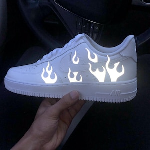 May include: White athletic shoe with reflective flame design. The shoe features a white sole and the word "AIR" on the side. The flame design is a bright, reflective white.