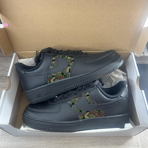 May include: A pair of black Air Force 1 sneakers with a camouflage pattern on the side panels, presented inside a cardboard box. The shoes have black laces and soles, with the "Air" logo visible on the side.
