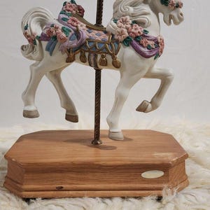 May include: A white carousel unicorn figurine with a golden horn, adorned with pink and purple floral accents. The unicorn stands on a wooden base, supported by a brass pole. The figurine is a decorative item.