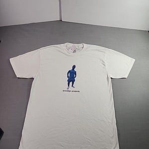 May include: White t-shirt with a blue graphic of a person and the text "brooklyn projects". The shirt is laid flat on a gray surface. The shirt has short sleeves and a crew neck.