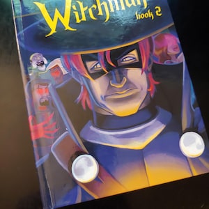 May include: A comic book titled "Witchman book 2" featuring a masked character with red hair and a blue and black outfit. The cover art has a dark background with yellow and white text.