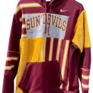 May include: Maroon and gold hoodie with the text "SUN DEVILS" across the chest. The hoodie features a kangaroo pocket, a hood, and a Nike logo. The design incorporates striped and textured panels.