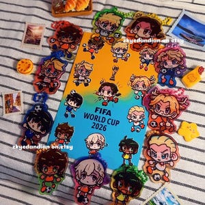 Könnte beinhalten: Eine Sammlung bunter Acryl-Schlüsselanhänger im Anime-Stil, die Fußballspieler darstellen, angeordnet um eine orange-blaue rechteckige Karte mit dem Text FIFA WORLD CUP 2026, ergänzt durch kleine dekorative Essens- und Landschaftsanhänger.